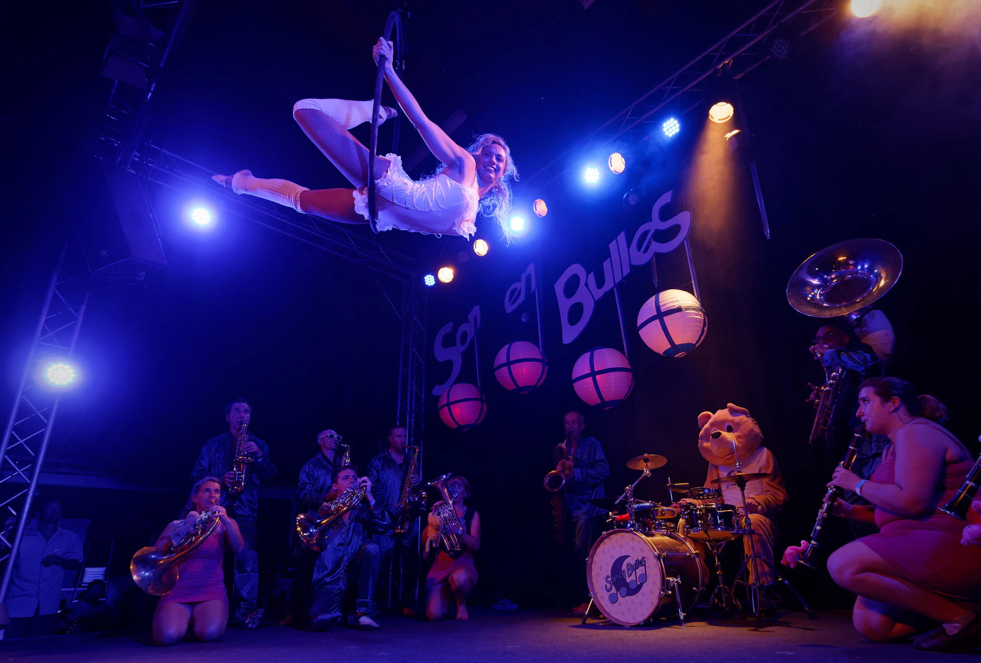 Aerial performer, live band and costumed character sharing one rehearsed circus scene on stage